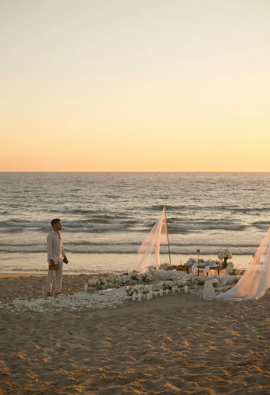 He waits on the beach at sunset beside a proposal set-up of linen sails, white flowers and candles